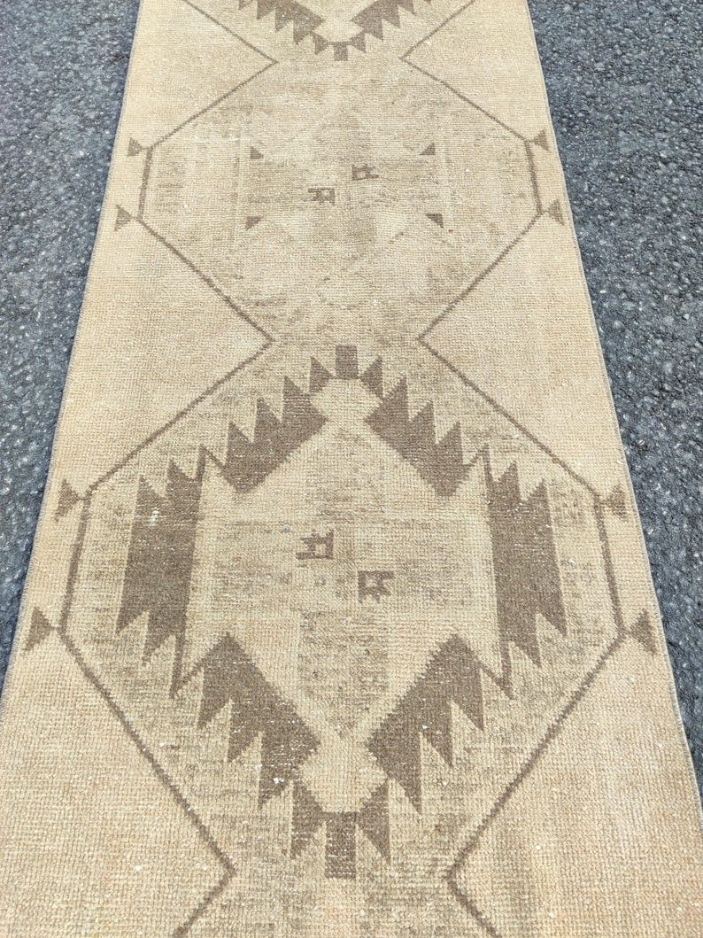 May include: Beige and brown patterned runner rug. The rug features a central diamond design with geometric shapes and a border of repeating patterns. The colour palette is neutral, with light beige and brown tones, creating a warm aesthetic.