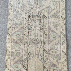 May include: A rectangular, vintage-style rug with a central design in beige, lavender, and teal. The rug features an intricate pattern of floral and geometric elements, with a detailed border. The rug's aged appearance suggests it is a handmade piece.