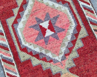 May include: A rectangular, hand-woven rug with a geometric pattern. The rug features a central diamond shape in shades of pink, grey, and white, surrounded by a red border. The rug's design includes star-like and zig-zag patterns.