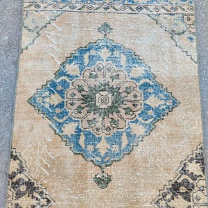 May include: A rectangular, vintage-style rug with a central diamond pattern in blue and green on a beige background. The rug features intricate border designs in blue and black, with a distressed, worn appearance.
