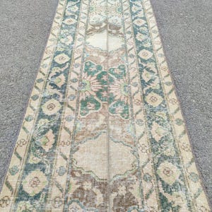Pode incluir: Um tapete retangular de estilo vintage com uma aparência desgastada. O tapete apresenta um campo central bege com padrões florais e geométricos em tons de verde, castanho e azul. As bordas têm uma borda mais escura.
