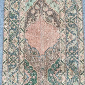 May include: A rectangular, vintage rug with a distressed appearance. The rug features a central design in shades of peach and brown, surrounded by borders in teal, green, and brown. The pattern includes floral and geometric elements.