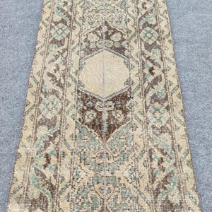 Op de afbeelding: Een rechthoekig vloerkleed in vintage stijl met een ingewikkeld, herhalend patroon. Het vloerkleed heeft een centraal ontwerp in beige, omgeven door bruine en blauwgroene accenten. Het kleurenpalet is gedempt, met een verweerde uitstraling.
