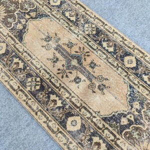 Può includere: Un tappeto rettangolare in stile vintage con un campo centrale beige e intricati motivi floreali blu scuro e marroni. Il tappeto ha un aspetto invecchiato, con un bordo con forme geometriche e motivi floreali.