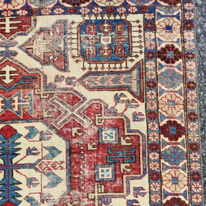 Puede incluir: Primer plano de una sección de una alfombra vintage con un patrón geométrico detallado. La alfombra presenta un campo central color crema con detalles en rojo y azul. El borde es azul oscuro con motivos florales y un borde exterior marrón.