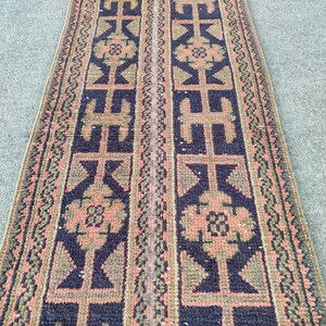 Könnte beinhalten: Ein rechteckiger Läuferteppich mit einem sich wiederholenden geometrischen Muster. Der Teppich hat einen marineblauen Hintergrund mit pfirsichfarbenen und olivgrünen Akzenten. Das Design umfasst stilisierte Formen und florale Motive.