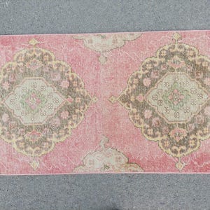 Könnte beinhalten: Ein rechteckiger Teppich im Vintage-Stil mit verblasstem rosa Hintergrund. Der Teppich weist sich wiederholende, verzierte Medaillonmuster in Braun-, Beige- und Grüntönen auf. Das Design des Teppichs deutet auf eine traditionelle oder böhmische Ästhetik hin.