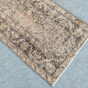 Può includere: Un tappeto rettangolare dall'aspetto invecchiato con un motivo floreale sbiadito nei toni del beige, marrone e verde oliva. Il design del tappeto presenta un campo centrale con un motivo ripetuto e un bordo. Il tappeto è posto su una superficie grigia.