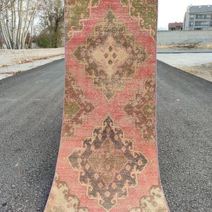 May include: A rectangular, vintage-style rug with a distressed appearance. The rug features a central medallion design in shades of brown, beige, and green, set against a faded red background. The rug is displayed outdoors on an asphalt surface.