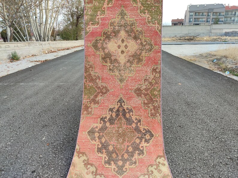 May include: A rectangular, vintage-style rug with a distressed appearance. The rug features a central medallion design in shades of brown, beige, and green, set against a faded red background. The rug is displayed outdoors on an asphalt surface.