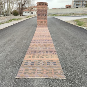 May include: A long, rectangular runner rug with a geometric pattern in shades of brown, orange, purple, and green. The rug is displayed outdoors on a paved surface, showcasing its length and design.