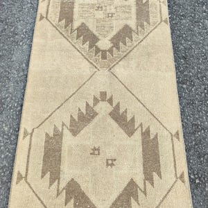 May include: A beige runner rug with a geometric pattern in shades of brown. The design features diamond shapes with stylized motifs. The rug is rectangular and appears to be made of woven material.