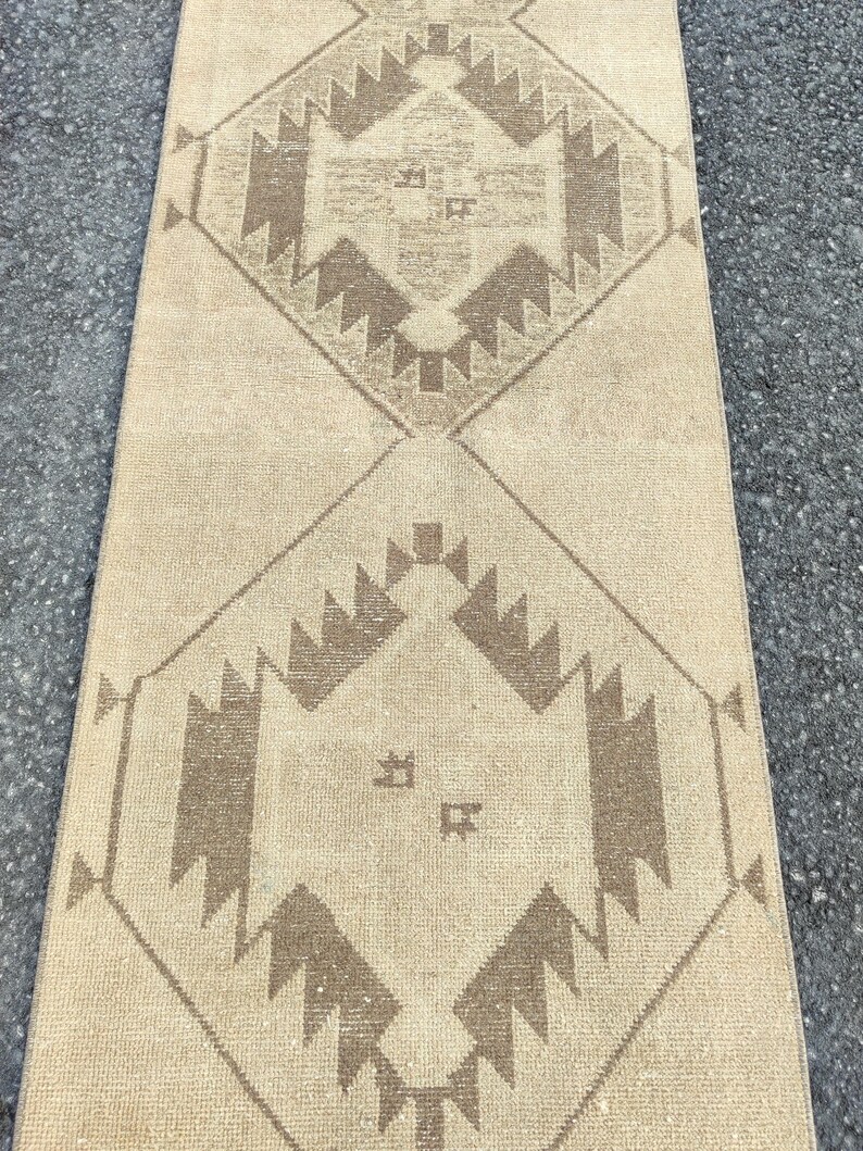 May include: A beige runner rug with a geometric pattern in shades of brown. The design features diamond shapes with stylized motifs. The rug is rectangular and appears to be made of woven material.