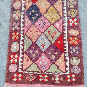 May include: A rectangular, hand-woven rug with a geometric pattern. The rug features a central diamond design in shades of pink, orange, and purple, framed by a border of red, white, and brown. The rug's design includes small tree-like motifs.