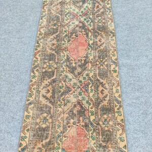 Könnte beinhalten: Ein langer, rechteckiger Teppich mit einem aufwendigen, verblichenen Muster. Der Teppich hat ein zentrales Design mit einer rötlich-orangenen Medaillonform, umgeben von einer Mischung aus braunen, beigen, grünen und blauen geometrischen und floralen Motiven. Die Ränder des Teppichs haben ein helleres, verwittertes Aussehen.