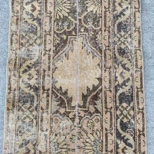 Op de afbeelding: Een rechthoekig vintage vloerkleed met een verweerde uitstraling. Het vloerkleed heeft een centraal beige veld met een symmetrisch patroon van bloemen en geometrische motieven in bruin, olijfgroen en zwart. De randen hebben een donkerdere rand.
