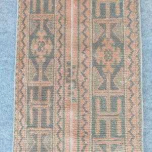 Puede incluir: Una alfombra rectangular de estilo vintage con un patrón geométrico repetitivo en tonos melocotón, gris y crema. La alfombra presenta una franja central de color melocotón flanqueada por diseños simétricos. La estética general recuerda a las alfombras orientales tradicionales.