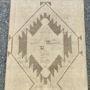 May include: A rectangular beige rug with a geometric pattern in brown tones. The central design is a diamond shape with stylized, angular motifs. The rug has a textured appearance and a fringed edge, suitable for home decor.