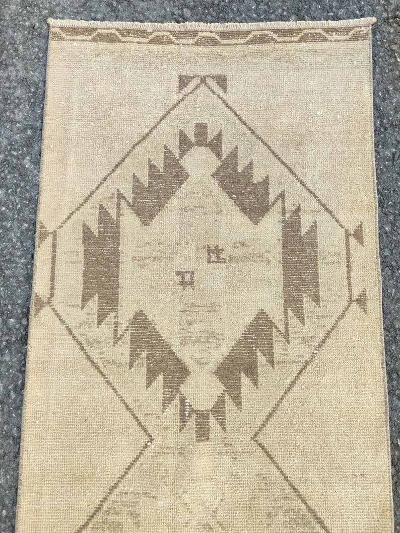 May include: A rectangular beige rug with a geometric pattern in brown tones. The central design is a diamond shape with stylized, angular motifs. The rug has a textured appearance and a fringed edge, suitable for home decor.