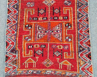 May include: A rectangular, hand-woven rug with a vibrant red field. The rug features intricate geometric patterns in orange, black, white, and brown. The border has a repeating zigzag design in various colours. The rug's design includes camel figures.