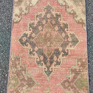 May include: A rectangular, distressed rug with a central medallion design. The rug features a faded colour palette of coral, brown, and olive green. The pattern includes geometric shapes and floral motifs, creating a vintage aesthetic.