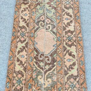May include: A vintage runner rug with a distressed appearance. The rug has a central design with a peach-coloured diamond shape, surrounded by intricate floral and geometric patterns in shades of brown, orange, green, and blue. The rug is rectangular and appears to be woven.