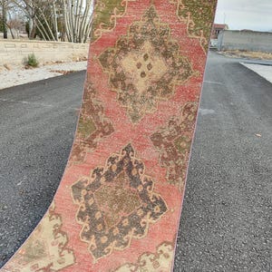 May include: A distressed runner rug with a faded appearance. The rug features a central medallion design in shades of brown, beige, and olive green, set against a faded red background. The rug is displayed outdoors on an asphalt surface.