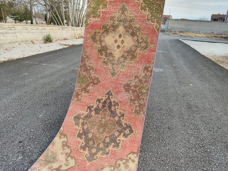 May include: A distressed runner rug with a faded appearance. The rug features a central medallion design in shades of brown, beige, and olive green, set against a faded red background. The rug is displayed outdoors on an asphalt surface.