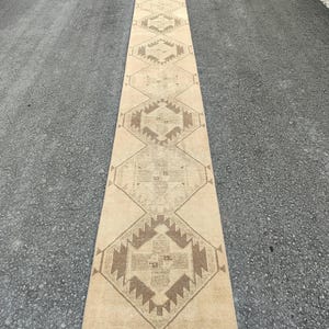 May include: A long, rectangular beige runner rug with a geometric pattern in shades of brown. The rug is placed on a dark asphalt surface, showcasing its length and design. The pattern features diamond and angular shapes.