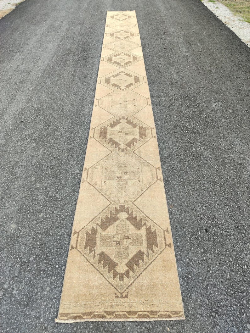 May include: A long, rectangular beige runner rug with a geometric pattern in shades of brown. The rug is placed on a dark asphalt surface, showcasing its length and design. The pattern features diamond and angular shapes.