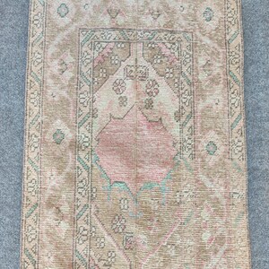 Peut inclure: Un tapis rectangulaire vintage à l'aspect délavé et vieilli. Le tapis présente un motif central dans des tons de rose et de crème, encadré par une bordure beige et turquoise. Le motif du tapis comprend des éléments floraux et géométriques.