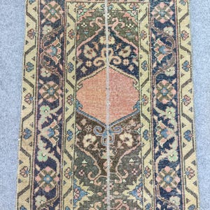 May include: A rectangular, vintage rug with an intricate floral pattern. The rug features a central design in shades of peach, olive green, and navy blue, framed by a border with similar colors and geometric motifs. The rug's dimensions are not visible.