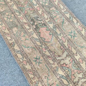 Könnte beinhalten: Vintage Läufer Teppich mit einem verblichenen Aussehen. Der Teppich zeigt ein sich wiederholendes Muster aus floralen und geometrischen Designs in gedämpften Rosa-, Grün- und Beigetönen. Der rechteckige Teppich ist aus gewebtem Material.