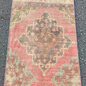 May include: A rectangular, vintage rug with a distressed look. It features a central medallion design in brown, grey, and pink, surrounded by floral and geometric patterns in muted green and blue tones. The rug's edges have a thin border. The rug is likely made of wool.