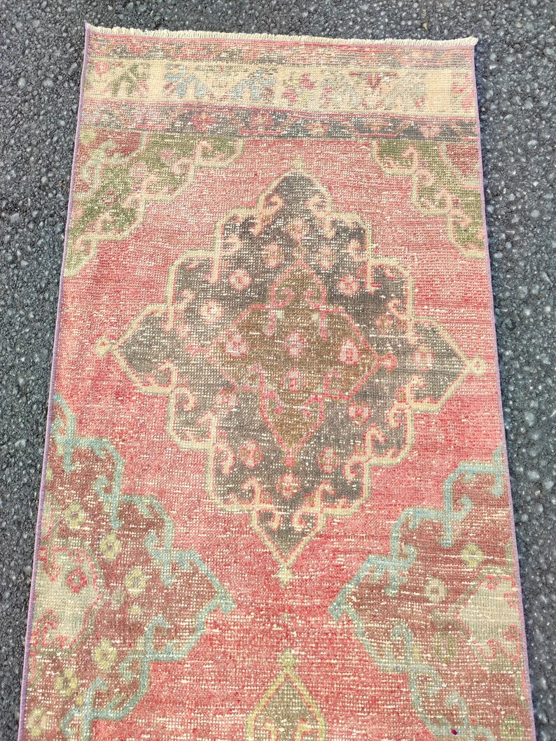 May include: A rectangular, vintage rug with a distressed look. It features a central medallion design in brown, grey, and pink, surrounded by floral and geometric patterns in muted green and blue tones. The rug's edges have a thin border. The rug is likely made of wool.