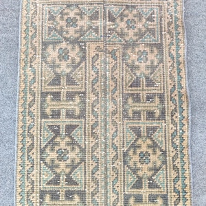 Peut inclure: Un tapis tissé rectangulaire présentant un motif géométrique dans des tons sourds de gris anthracite, vert sauge et beige. Le design comprend des motifs symétriques et une colonne centrale verticale, avec une courte frange brun-rouge au sommet.