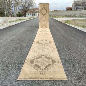 May include: A long, beige runner rug featuring geometric patterns in brown hues. The rug is displayed on a paved road, highlighting its length and design. The intricate details and neutral colours of the rug make it a versatile home decor item, suitable for various spaces.