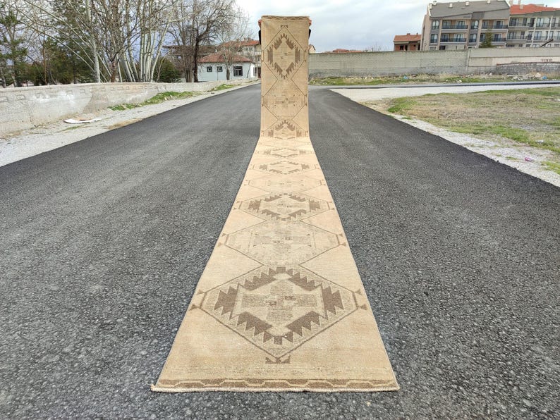 May include: A long, beige runner rug featuring geometric patterns in brown hues. The rug is displayed on a paved road, highlighting its length and design. The intricate details and neutral colours of the rug make it a versatile home decor item, suitable for various spaces.