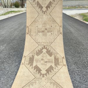 May include: A beige runner rug with a geometric pattern in shades of brown. The rug is rectangular with a central design of diamonds and zigzags. The rug is displayed outdoors on a paved surface.