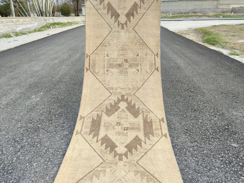 May include: A beige runner rug with a geometric pattern in shades of brown. The rug is rectangular with a central design of diamonds and zigzags. The rug is displayed outdoors on a paved surface.