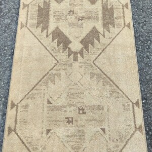May include: A beige and brown patterned rug. The rug has a repeating geometric design with diamond and triangular shapes. The colour palette is neutral, with a beige background and brown accents. The rug is rectangular and appears to be made of woven material.