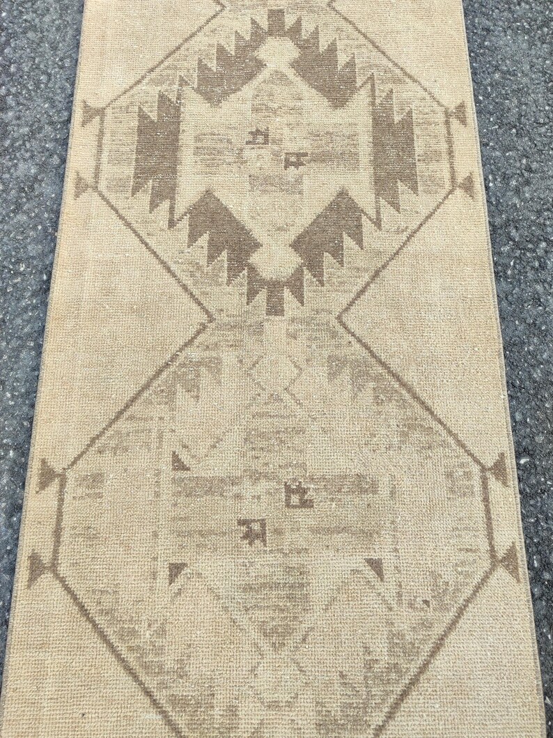 May include: A beige and brown patterned rug. The rug has a repeating geometric design with diamond and triangular shapes. The colour palette is neutral, with a beige background and brown accents. The rug is rectangular and appears to be made of woven material.