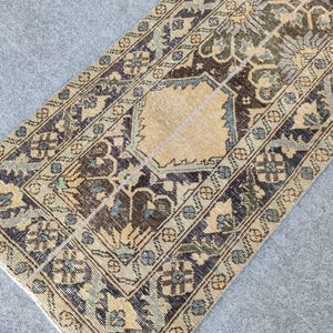 Peut inclure: Un tapis de couloir ancien avec un médaillon central beige et des motifs floraux complexes en marine, marron et vert olive. La bordure du tapis présente un motif répétitif, ajoutant à son esthétique vintage. Le tapis est rectangulaire.