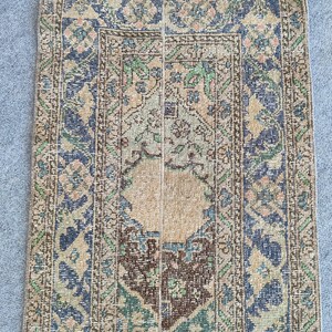 Könnte beinhalten: Ein antiker, rechteckiger Teppich mit einem verwitterten, Vintage-Aussehen. Der Teppich hat ein zentrales beiges Feld, das von aufwendigen Bordüren in Blau-, Grün- und Brauntönen umrahmt wird. Florale und geometrische Muster zieren die Oberfläche des Teppichs und schaffen eine traditionelle Ästhetik.