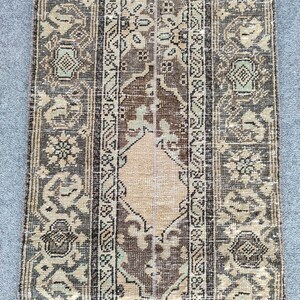 Peut inclure: Un tapis rectangulaire de style vintage avec un panneau central marron et des bordures à motifs. Le tapis présente un motif en forme de losange beige au centre, entouré de motifs floraux et géométriques dans des tons de marron, beige et vert. Le tapis a un aspect vieilli.