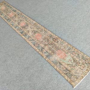 Peut inclure: Un long tapis rectangulaire d'aspect vieilli. Il présente des motifs floraux délavés en pêche, sarcelle et vert sur un fond beige. Les bords sont finis avec une bordure subtile. Idéal pour un couloir.