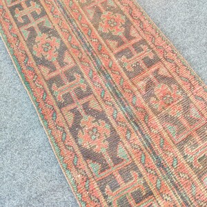Peut inclure: Tapis rectangulaire à motifs géométriques répétés, dans des tons de marron, corail et turquoise. Le tapis présente un aspect vieilli, avec des bords légèrement effilochés. Le motif suggère un style traditionnel. Le tapis est probablement en laine ou dans une matière similaire.