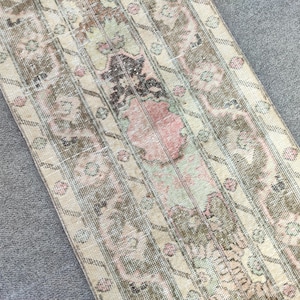Könnte beinhalten: Ein rechteckiger Teppich im Vintage-Stil mit verblasstem, abgenutztem Aussehen. Der Teppich weist ein sich wiederholendes Muster aus floralen und geometrischen Elementen in gedämpften Tönen von Beige, Braun, Rosa und Grün auf. Der Teppich liegt auf einer grauen Oberfläche.
