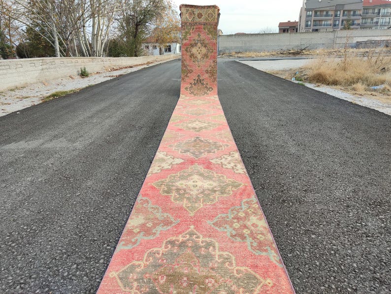 May include: A long, rectangular rug with a detailed pattern in red, beige, and green. The rug is laid out on a dark asphalt surface, highlighting its intricate design and colour palette.