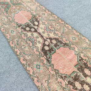 May include: A vintage runner rug with a distressed, faded appearance. The rug features a central design in shades of pink and brown, framed by borders in similar tones with accents of green and cream. The rug's design is intricate and detailed.